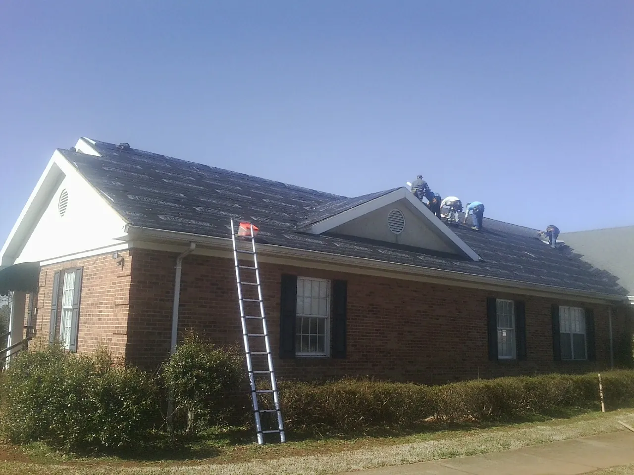 Crew tearing shingles off a brick ranch roof with a ladder set — Metal Roof Repair work by our crew in Milton